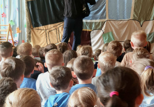 Na zdjęciu widać scenę, na której występuje aktor ubrany na czarno. Ma na głowie czapkę oraz okulary, co nadaje mu charakterystyczny wygląd sceniczny. Aktor odgrywa swoją rolę, zwracając się w stronę publiczności lub innych postaci. W tle widoczna jest dekoracja sceniczna dopasowana do tematyki przedstawienia. Przed sceną siedzą dzieci z przedszkola, które z zainteresowaniem oglądają spektakl i skupiają uwagę na występie.