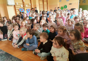 Na zdjęciu widać dzieci z przedszkola siedzące na podłodze i oglądające teatrzyk pt. „Wiosenne porządki”. Przed nimi występuje dwoje aktorów na tle scenografii przedstawiającej dom. Aktorzy posługują się rekwizytami i odgrywają scenkę. Dzieci są skupione i z zainteresowaniem śledzą przebieg przedstawienia. W tle widoczny jest napis „Witaj Wiosno” oraz kolorowe dekoracje o tematyce wiosennej.