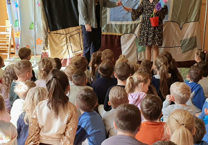 Na zdjęciu widać dzieci z przedszkola siedzące na podłodze i oglądające przedstawienie teatralne. Przed nimi stoi dwoje aktorów na tle dekoracji przypominającej dom. Jedna z osób trzyma rekwizyty, a druga mówi swoją kwestię. Dzieci są skupione i uważnie obserwują spektakl. W tle widoczny jest napis „Witaj Wiosno” oraz kolorowe dekoracje nawiązujące do wiosny.