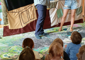 Na zdjęciu widać dwoje aktorów – kobietę i mężczyznę – którzy występują na scenie i wcielają się w swoje role w spektaklu. Aktorzy stoją naprzeciw siebie i odgrywają scenkę z wykorzystaniem rekwizytów. Ich gesty i mimika wskazują na zaangażowanie w przedstawienie. W tle widoczna jest dekoracja sceniczna, a przed sceną siedzą dzieci, które z uwagą oglądają występ.