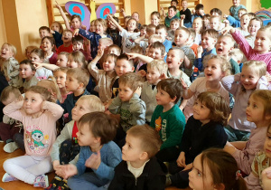 Na zdjęciu widać grupę dzieci siedzących na podłodze w sali przedszkolnej, które oglądają przedstawienie teatralne. Na scenie występują aktorzy w kolorowych strojach, odgrywając scenki z wykorzystaniem rekwizytów. Dzieci są zwrócone w stronę sceny, siedzą blisko siebie i z dużym zainteresowaniem obserwują przebieg spektaklu.