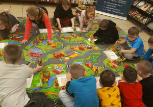 Na zdjęciu widać dzieci siedzące na dywanie w Bibliotece Pedagogicznej. Przedszkolaki rysują swoje ilustracje związane z książkami, korzystając z kartek i kredek. Dzieci są skupione na wykonywanych zadaniach, a wokół nich widoczne są materiały plastyczne. W tle znajdują się regały z książkami.
