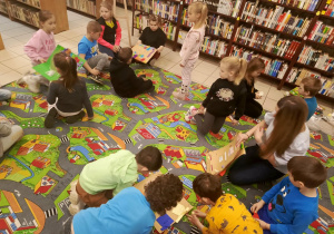 Na zdjęciu przedstawiono dzieci pracujące na dywanie podczas zajęć w Bibliotece Pedagogicznej. Przedszkolaki wykonują zadania związane z tematyką książek. Widać ich zaangażowanie i skupienie. W tle widoczne są półki z książkami oraz przestrzeń biblioteczna.