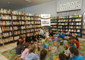 Na zdjęciu widać dzieci z grupy przedszkolnej siedzące na kolorowym dywanie. Przed nimi i wokół nich znajdują się rozłożone książki, które dzieci oglądają z zainteresowaniem. W tle widoczne są regały wypełnione książkami oraz napis „Biblioteka Pedagogiczna”. Dzieci są skupione i aktywnie uczestniczą w zajęciach czytelniczych.