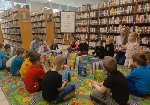 Na zdjęciu przedstawiono dzieci siedzące na dywanie w otoczeniu książek. Przedszkolaki przeglądają wybrane pozycje i rozmawiają między sobą. W tle znajdują się wysokie regały z książkami oraz widoczny napis „Biblioteka Pedagogiczna”. Atmosfera jest spokojna i sprzyja rozwijaniu zainteresowań czytelniczych dzieci.
