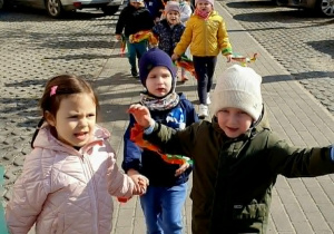 Na zdjęciu przedstawiono fragment pochodu – dzieci idą w grupie, machając kolorowymi ozdobami. Słońce oświetla scenę, a w tle widoczne są ogrodzenie, drzewa i budynki mieszkalne. Atmosfera jest radosna i pełna energii.