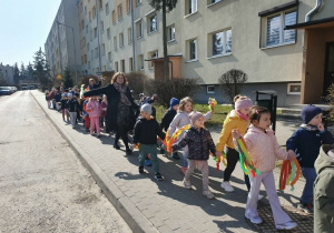 Na zdjęciu widoczna jest grupa dzieci maszerujących chodnikiem w słoneczny dzień. Dzieci trzymają w rękach kolorowe wstążki i unoszą je do góry. Na czele grupy idzie nauczycielka, prowadząc pochód. W tle znajdują się drzewa, ogrodzenie oraz zabudowania mieszkalne.