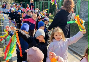 Na zdjęciu widać grupę małych dzieci z przedszkola podczas spaceru. Idą chodnikiem wzdłuż ogrodzenia, prowadzone przez nauczycielkę znajdującą się na początku grupy. Dzieci są ubrane w kurtki i czapki, a w rękach trzymają kolorowe wstążki, którymi machają. Panuje słoneczna pogoda. W tle widoczne są drzewa oraz budynki mieszkalne. Dzieci wyglądają na radosne i zaangażowane w wspólny marsz.
