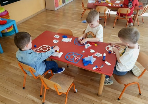 Dzieci pracują w skupieniu, rozwijając swoją kreatywność oraz sprawność manualną. W tle widać salę przedszkolną z kolorowym dywanem i wyposażeniem dostosowanym do potrzeb najmłodszych. Atmosfera jest radosna i sprzyja twórczej zabawie.