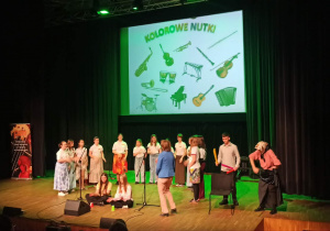 Na zdjęciu widoczna jest scena podczas koncertu muzycznego. Na scenie stoją uczniowie szkoły muzycznej, którzy prezentują swoje umiejętności wokalne i instrumentalne. Część z nich śpiewa przy mikrofonach, inni wykonują rytmiczne układy z wykorzystaniem prostych instrumentów. W tle znajduje się duży ekran z napisem „Kolorowe nutki” oraz ilustracjami różnych instrumentów muzycznych. Scena oświetlona jest kolorowym światłem, które podkreśla artystyczny charakter wydarzenia. Występ odbywa się przed zgromadzoną publicznością, a całość ma radosny i edukacyjny charakter, przybliżając dzieciom świat muzyki i różnorodność instrumentów.