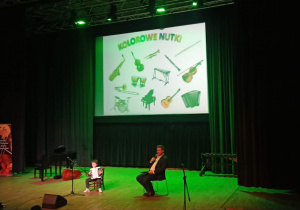 Na zdjęciu widoczny jest uczeń stojący na scenie, który prezentuje grę na instrumencie. Dziecko skupione jest na wykonaniu utworu, a obok znajduje się mikrofon. W tle widać elementy scenografii oraz ekran.