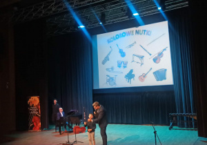 Na zdjęciu uczeń prezentuje instrument, pokazując jego budowę oraz sposób wydobywania dźwięku. Dzieci z widowni słuchają z zainteresowaniem, poznając nowe brzmienia.