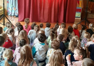Dzieci siedzą na dywanie w sali przedszkolnej, zwrócone w stronę sceny. Z uwagą i zaciekawieniem oglądają przedstawienie przygotowane przez aktorów.