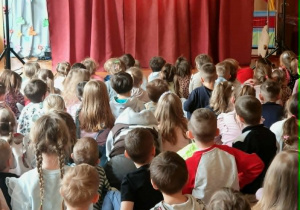 Dzieci oglądają spektakl teatralny w przedszkolu. W tle widoczna jest scenografia oraz aktorzy w kolorowych kostiumach. Dzieci są zaciekawione i zaangażowane.