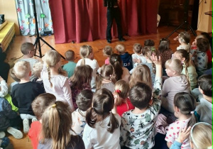 Grupa dzieci siedzi na dywanie i patrzy na scenę. Panuje cisza i skupienie, a przedstawienie wzbudza duże zainteresowanie wśród przedszkolaków.