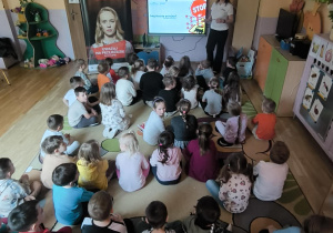 Zdjęcie przedstawia grupę przedszkolaków uczestniczących w zajęciach edukacyjnych o bezpieczeństwie na torach. Dzieci siedzą na dywanie w kolorowej sali i wspólnie poznają zasady bezpiecznego zachowania w pobliżu kolei.