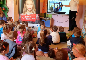 Zdjęcie przedstawia przedszkolaki podczas oglądania prezentację o bezpieczeństwie na torach. Dzieci skupiają uwagę na ekranie multimedialnym, aktywnie uczestnicząc w zajęciach profilaktycznych.