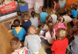 Na zdjęciu dzieci siedzą w sali przedszkolnej i oglądają prezentację dotyczącą zasad bezpieczeństwa na przejazdach kolejowych. Przedszkolaki z zainteresowaniem słuchają prowadzącej zajęcia nauczycielki oraz oglądają materiały wyświetlane na ekranie multimedialnym.