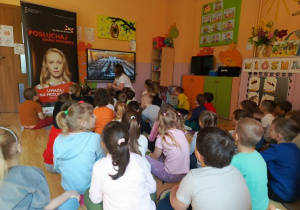 Na zdjęciu widać grupę dzieci siedzących na podłodze w sali przedszkolnej i uczestniczących w zajęciach edukacyjnych dotyczących bezpieczeństwa na torach kolejowych. Nauczycielka prowadzi prezentację wyświetlaną na ekranie multimedialnym. Obok ekranu stoi baner informacyjny związany z akcją „Bezpieczny przejazd”. Sala jest kolorowa i udekorowana pracami dzieci oraz wiosennymi dekoracjami. Dzieci uważnie słuchają i obserwują prezentowane materiały edukacyjne.