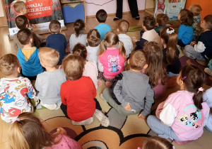 Zdjęcie przedstawia grupę przedszkolaków uczestniczących w zajęciach edukacyjnych o bezpieczeństwie na torach. Dzieci siedzą na dywanie w kolorowej sali i wspólnie poznają zasady bezpiecznego zachowania w pobliżu kolei.