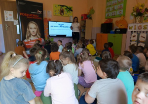 Na fotografii widać dzieci biorące udział w akcji edukacyjnej dotyczącej bezpieczeństwa kolejowego. W tle znajduje się baner kampanii „Bezpieczny przejazd”, a nauczycielka omawia z dziećmi ważne zasady związane z ruchem kolejowym.