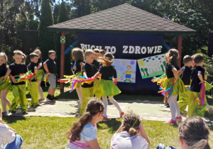 Na zdjęciu widoczna jest grupa dzieci prezentujących występ taneczny na terenie przedszkola. Dzieci ubrane są w jednakowe stroje – czarne koszulki oraz zielone spodnie lub spódniczki. W dłoniach trzymają kolorowe pompony w odcieniach niebieskiego, żółtego, zielonego, różowego i czerwonego. Przedszkolaki wykonują układ choreograficzny przed altaną znajdującą się na placu przedszkolnym. W tle widoczny jest baner związany z wydarzeniem oraz otaczająca teren zieleń. Na pierwszym planie znajdują się dzieci i osoby dorosłe oglądające występ. Zdjęcie przedstawia część artystyczną wydarzenia zorganizowanego w ramach rozstrzygnięcia konkursu plastycznego „Ruch to zdrowie”. Atmosfera jest radosna i pełna energii, a występ podkreśla znaczenie aktywności ruchowej i wspólnej zabawy.