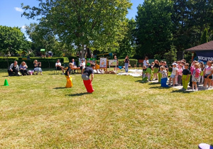 Na zdjęciu widać dzieci uczestniczące w konkurencji sportowej na terenie przedszkolnego placu. W centralnej części fotografii dwoje dzieci bierze udział w wyścigu w workach na trawiastej powierzchni. Po prawej stronie stoi grupa dzieci oczekujących na swoją kolej i obserwujących przebieg zabawy. W tle znajdują się nauczyciele, opiekunowie oraz goście siedzący na krzesłach. Widoczne są także tablice z pracami konkursowymi oraz elementy dekoracyjne związane z wydarzeniem. Po prawej stronie częściowo widoczna jest altana z banerem promującym konkurs „Ruch to zdrowie”. Całość odbywa się na świeżym powietrzu, w słoneczny dzień, w otoczeniu zieleni i wysokich drzew. Zdjęcie przedstawia aktywną zabawę ruchową podczas festynu i rozstrzygnięcia konkursu plastycznego „Ruch to zdrowie”.