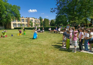 Na zdjęciu dzieci uczestniczą w konkurencjach sportowych na świeżym powietrzu. Przedszkolaki wykonują zadania ruchowe przygotowane przez organizatorów. Wydarzenie odbywa się na trawniku przedszkolnym w otoczeniu zieleni.