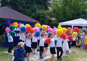 Grupa dzieci przedszkolnych prezentuje układ taneczny podczas Festynu Rodzinnego. Dzieci ubrane są w jasne stroje i trzymają kolorowe pompony, którymi wykonują elementy choreografii. Występ odbywa się na świeżym powietrzu, a zgromadzeni rodzice i goście obserwują pokaz. Na twarzach dzieci widoczne jest zaangażowanie i radość ze wspólnego występu. Kolorowe rekwizyty podkreślają uroczysty i rodzinny charakter wydarzenia.
