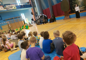 Przedszkolaki siedzą na widowni, ułożone w rzędach na krzesłach, zwrócone w stronę sceny. Z dużym zainteresowaniem i skupieniem oglądają spektakl przygotowany przez starszych uczniów szkoły. Na ich twarzach widać zaciekawienie oraz zaangażowanie w oglądaną historię.