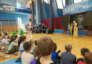 Przedszkolaki siedzą na widowni, zwrócone w stronę sceny. Z dużym zainteresowaniem i skupieniem oglądają spektakl przygotowany przez starszych uczniów szkoły. Na ich twarzach widać zaciekawienie oraz zaangażowanie w oglądaną historię.