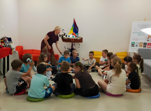 W sali bibliotecznej dzieci siedzą w kręgu na kolorowych poduszkach i uczestniczą w spotkaniu z autorką książek dla dzieci. Prowadząca rozmawia z przedszkolakami oraz prezentuje rekwizyty zw