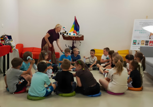 W sali bibliotecznej dzieci siedzą w kręgu na kolorowych poduszkach i uczestniczą w spotkaniu z autorką książek dla dzieci. Prowadząca rozmawia z przedszkolakami oraz prezentuje rekwizyty związane z tematyką książek. Dzieci z zainteresowaniem słuchają i aktywnie biorą udział w zajęciach. W tle widoczne są kolorowe meble, dekoracje oraz ekspozycja promująca twórczość autorki. Spotkanie odbywa się w przyjaznej atmosferze, sprzyjającej rozwijaniu zainteresowań czytelniczych najmłodszych.