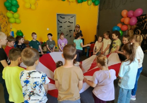 Grupa przedszkolaków stoi w kręgu i uczestniczy we wspólnej zabawie z chustą animacyjną. Dzieci trzymają kolorową chustę i wykonują ćwiczenia ruchowe pod kierunkiem prowadzącego. Zajęcia odbywają się w sali udekorowanej kolorowymi balonami. Na twarzach uczestników widać zaangażowanie i radość ze wspólnej zabawy. Aktywność sprzyja integracji grupy oraz rozwijaniu sprawności ruchowej i współpracy.