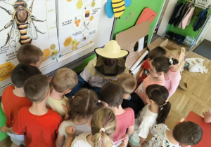 Na zdjęciu widać grupę dzieci zgromadzonych wokół osoby prezentującej strój pszczelarza podczas zajęć edukacyjnych w sali przedszkolnej. Dzieci z dużym zainteresowaniem obserwują elementy wyposażenia pszczelarza, w tym kapelusz z ochronną siatką. W tle znajduje się tablica z dużymi planszami przedstawiającymi budowę pszczoły oraz informacje dotyczące życia tych owadów. Sala jest kolorowa i wyposażona w pomoce dydaktyczne związane z tematyką pszczół. Spotkanie ma charakter praktyczny i umożliwia dzieciom bezpośrednie poznawanie pracy pszczelarza.
