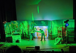 Na zdjęciu widoczna jest scena teatralna podczas przedstawienia „Karolcia” w Centrum Integracji Ratusz. Na scenie znajduje się troje aktorów odgrywających jedną ze scen spektaklu. W tle ustawiona jest kolorowa scenografia przedstawiająca miejskie otoczenie, a po bokach sceny widoczne są elementy dekoracji i oświetlenia. W dolnej części zdjęcia można zauważyć publiczność zgromadzoną na widowni, wśród której znajdują się dzieci z przedszkola. Przedstawienie dostarcza młodym widzom wielu emocji oraz pozwala rozwijać zainteresowania teatralne i kulturalne.