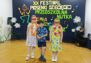 Troje dzieci stoi na tle kolorowej dekoracji przygotowanej na wydarzenie. Dzieci są ubrane odświętnie i ustawione obok siebie, zwrócone w stronę aparatu. W tle widoczne są elementy scenografii nawiązujące do festiwalu piosenki, które tworzą radosną, uroczystą atmosferę.