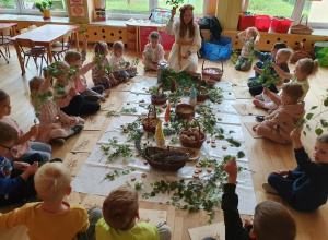 Na zdjęciu dzieci z grupy przedszkolnej siedzą w kręgu na podłodze wokół kompozycji ułożonej z gałązek, ziół, kwiatów i naturalnych materiałów. W środku siedzi prowadząca warsztaty, która pr