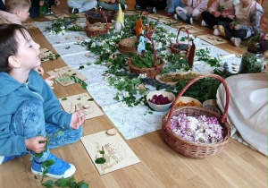Dzieci wspólnie uczestniczą w zajęciach przyrodniczo-plastycznych. Na środku sali znajdują się koszyki z ziołami, kwiatami i liśćmi, które przedszkolaki oglądają i wykorzystują podczas twórczej pracy.