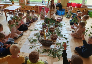 Na zdjęciu dzieci z grupy przedszkolnej siedzą w kręgu na podłodze wokół kompozycji ułożonej z gałązek, ziół, kwiatów i naturalnych materiałów. W środku siedzi prowadząca warsztaty, która prezentuje dzieciom rośliny i opowiada o ich właściwościach. Przedszkolaki z zainteresowaniem unoszą gałązki i zioła, aktywnie uczestnicząc w zajęciach. Warsztaty odbywają się w jasnej sali przedszkolnej, a na podłodze rozłożone są koszyki z roślinami oraz materiały wykorzystywane podczas zajęć. Atmosfera spotkania jest spokojna, twórcza i pełna zaangażowania dzieci.