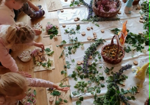 Przedszkolaki z zaangażowaniem układają naturalne materiały na drewnianych sercach. Na stolikach znajdują się przygotowane rośliny, klej oraz elementy dekoracyjne wykorzystywane podczas warsztatów.