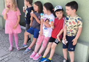 Na zdjęciu sześcioro dzieci stoi i siedzi przy ścianie budynku podczas wyjścia poza teren przedszkola. Przedszkolaki trzymają w dłoniach lody w wafelkach i z uśmiechem delektują się letnim poczęstunkiem. Dzieci ubrane są w kolorowe, lekkie stroje odpowiednie na ciepłą pogodę. W tle widoczna jest ściana budynku oraz fragment chodnika. Zdjęcie przedstawia radosny moment wspólnego odpoczynku po spacerze.