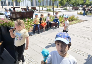 Grupa dzieci spożywa lody podczas wyjścia poza teren przedszkola. Wspólny poczęstunek jest okazją do integracji oraz miłego spędzenia czasu na świeżym powietrzu.