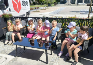 Dzieci z grupy przedszkolnej siedzą przy stoliku na świeżym powietrzu i jedzą lody. Przedszkolaki rozmawiają ze sobą i cieszą się wspólnie spędzonym czasem podczas letniego spaceru.