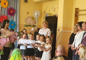 Rodzice podczas uroczystości zakończenia roku przedszkolnego wręczają kwiaty i podziękowania nauczycielom oraz pracownikom przedszkola. W tle widoczne są kolorowe dekoracje i okolicznościowa scenografia.