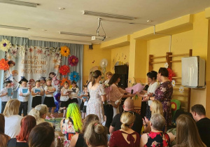 Podczas uroczystości zakończenia roku przedszkolnego rodzice wręczają kwiaty i podziękowania nauczycielom oraz pracownikom przedszkola. Na środku sali stoi grupa przedszkolaków ubranych odświętnie, trzymających pamiątkowe upominki. Obok znajdują się nauczyciele i pracownicy placówki, a jedna z osób przemawia do zgromadzonych gości. W tle widoczne są kolorowe dekoracje kwiatowe oraz okolicznościowa scenografia związana z zakończeniem edukacji przedszkolnej. Na pierwszym planie siedzą rodzice i zaproszeni goście uczestniczący w uroczystości.