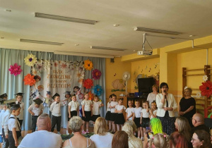 Przedszkolaki podczas występu z okazji zakończenia roku przedszkolnego. Dzieci prezentują program artystyczny przed rodzicami, nauczycielami i zaproszonymi gośćmi, którzy nagradzają ich brawami.