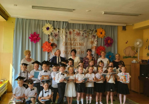 Pamiątkowe zdjęcie przedstawia dzieci kończące edukację przedszkolną wraz z nauczycielami i zaproszonym gościem. Część dzieci ma na głowach birety, a w dłoniach pamiątkowe upominki. Sala udekorowana jest dużymi papierowymi kwiatami i okolicznościowym banerem.
