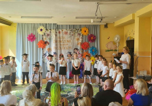 Przedszkolaki podczas występu artystycznego z okazji zakończenia przedszkola. Dzieci prezentują program słowno-muzyczny na tle kolorowej dekoracji i okolicznościowego napisu.