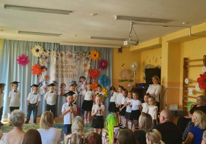 Dzieci z grupy starszaków stoją na scenie podczas występu artystycznego z okazji zakończenia przedszkola. Przedszkolaki recytują wiersze i śpiewają piosenki przed zgromadzonymi rodzicami oraz gośćmi.